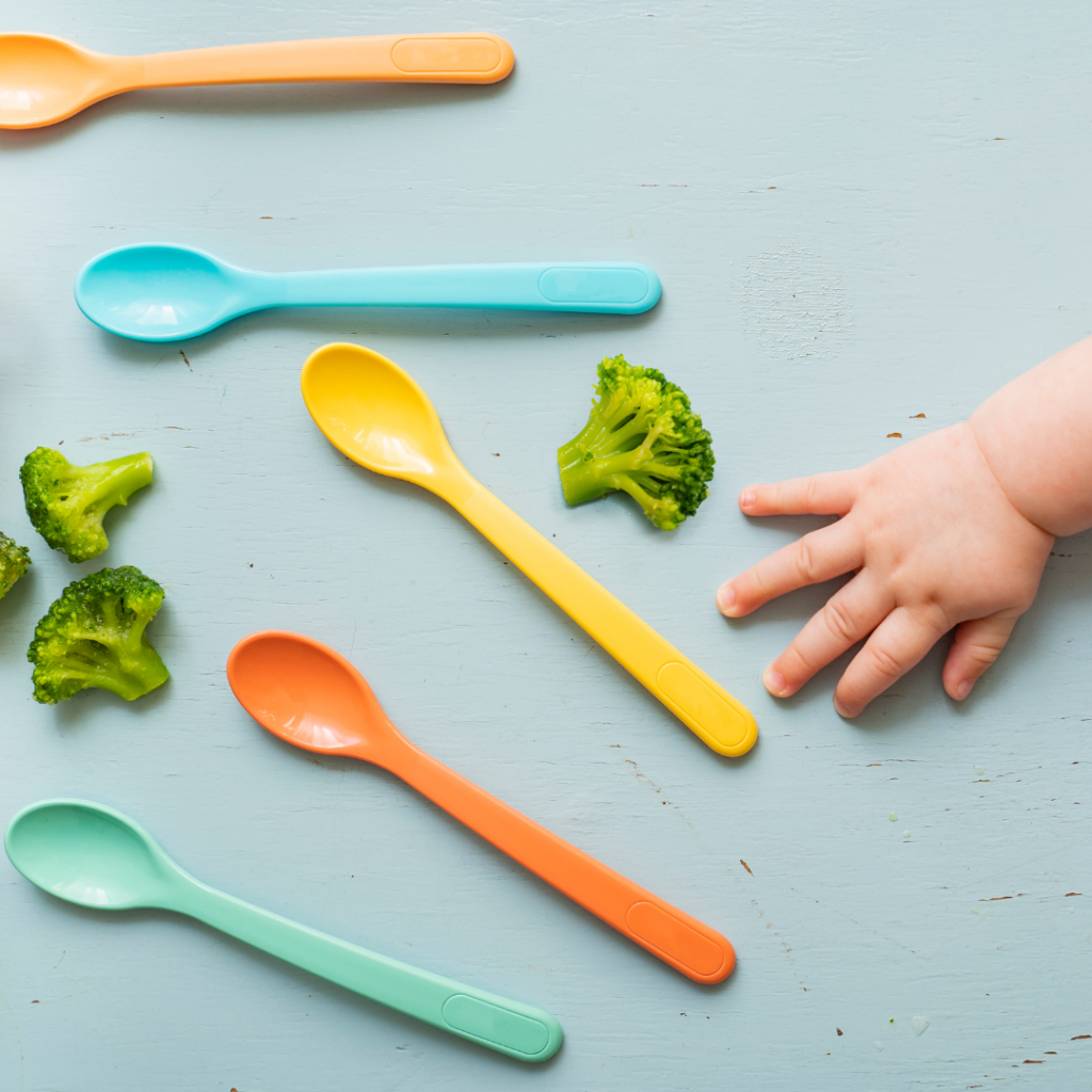 Colorful plastic spoons for feeding babies and broccoli florets on a table, with a baby's hand reaching towards them.