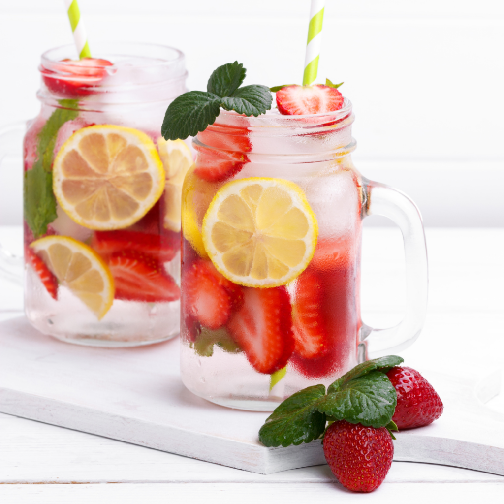 Two glass mason jars filled with water, strawberries, lemon, and ice cubes with green-stripped straws. 