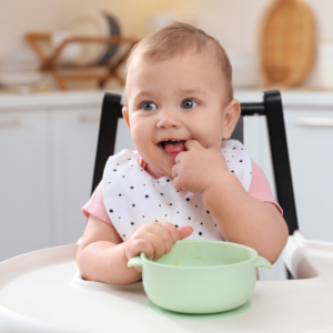 Baby smiling while sitting in a high chair with a bowl of baby food in front of them.