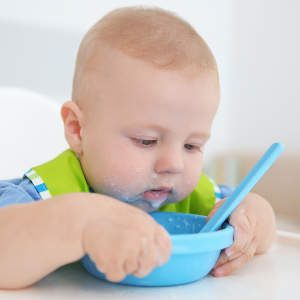 Baby in a high chair holding a blue bowl, eating a solid meal.