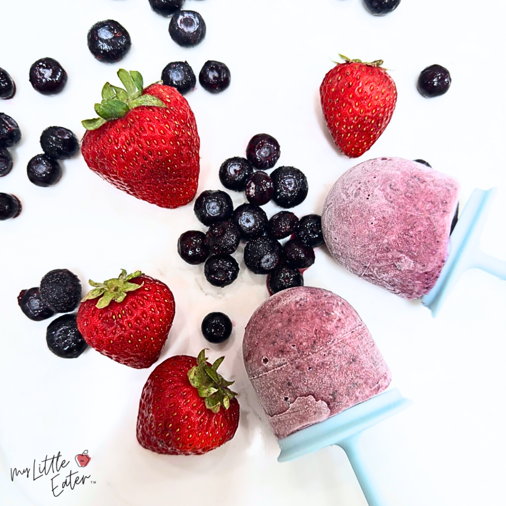 Two deep red popsicles with strawberries and blueberries beside.