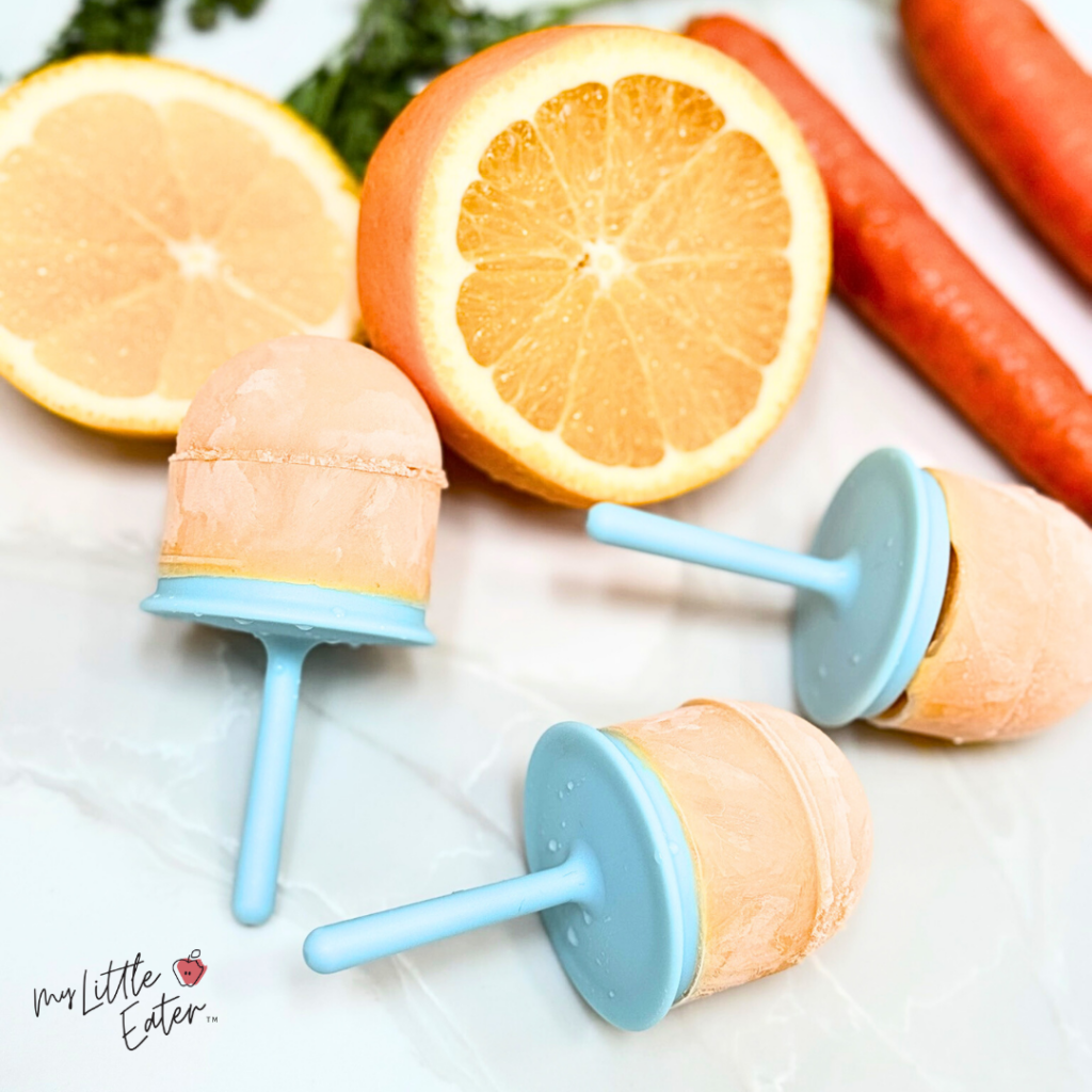 Three orange popsicles with oranges and carrots in the background.