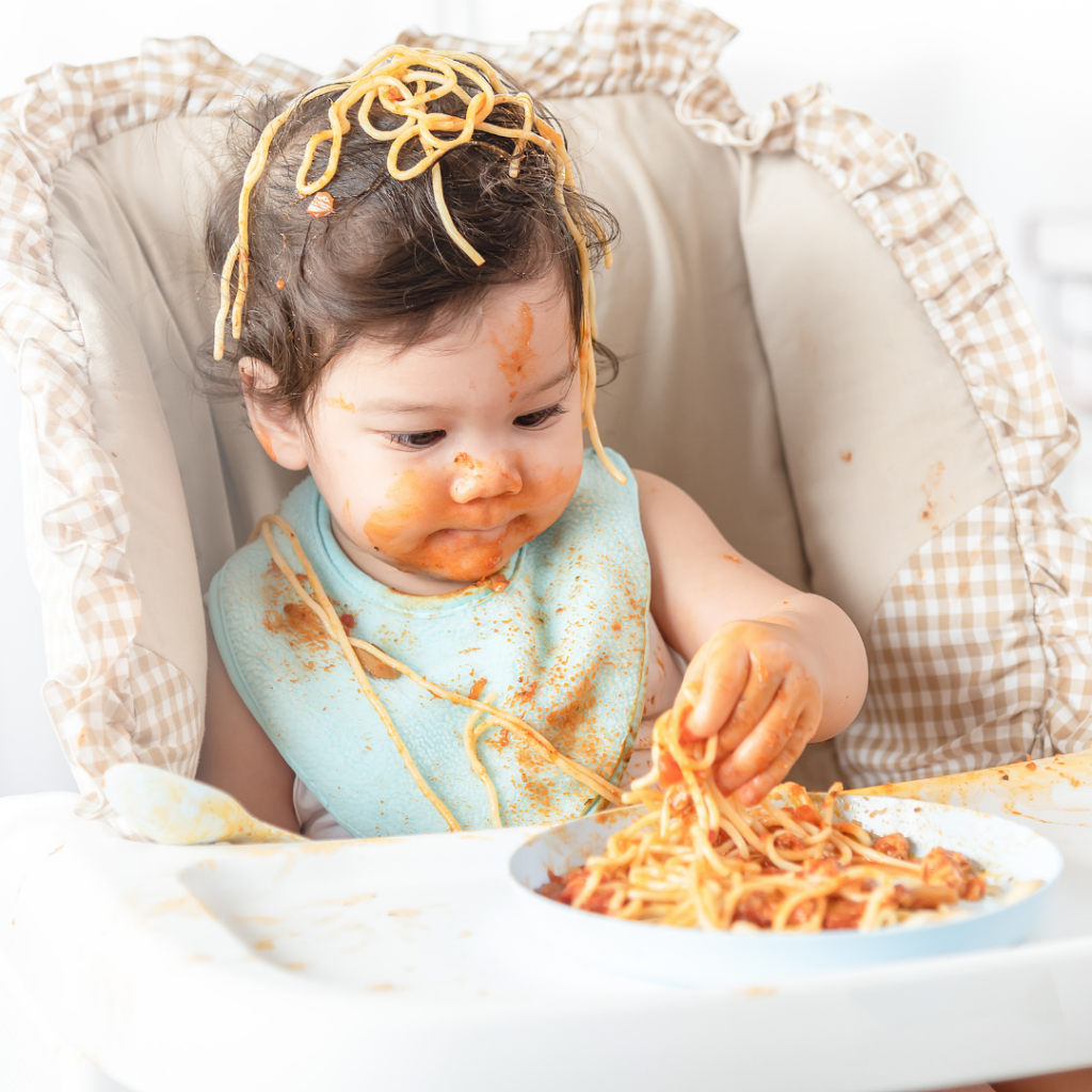 Baby of average weight eats spaghetti in a high chair.