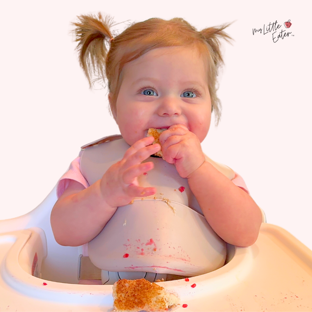 Baby sitting in her high chair eating toast strips topped with mashed berries.