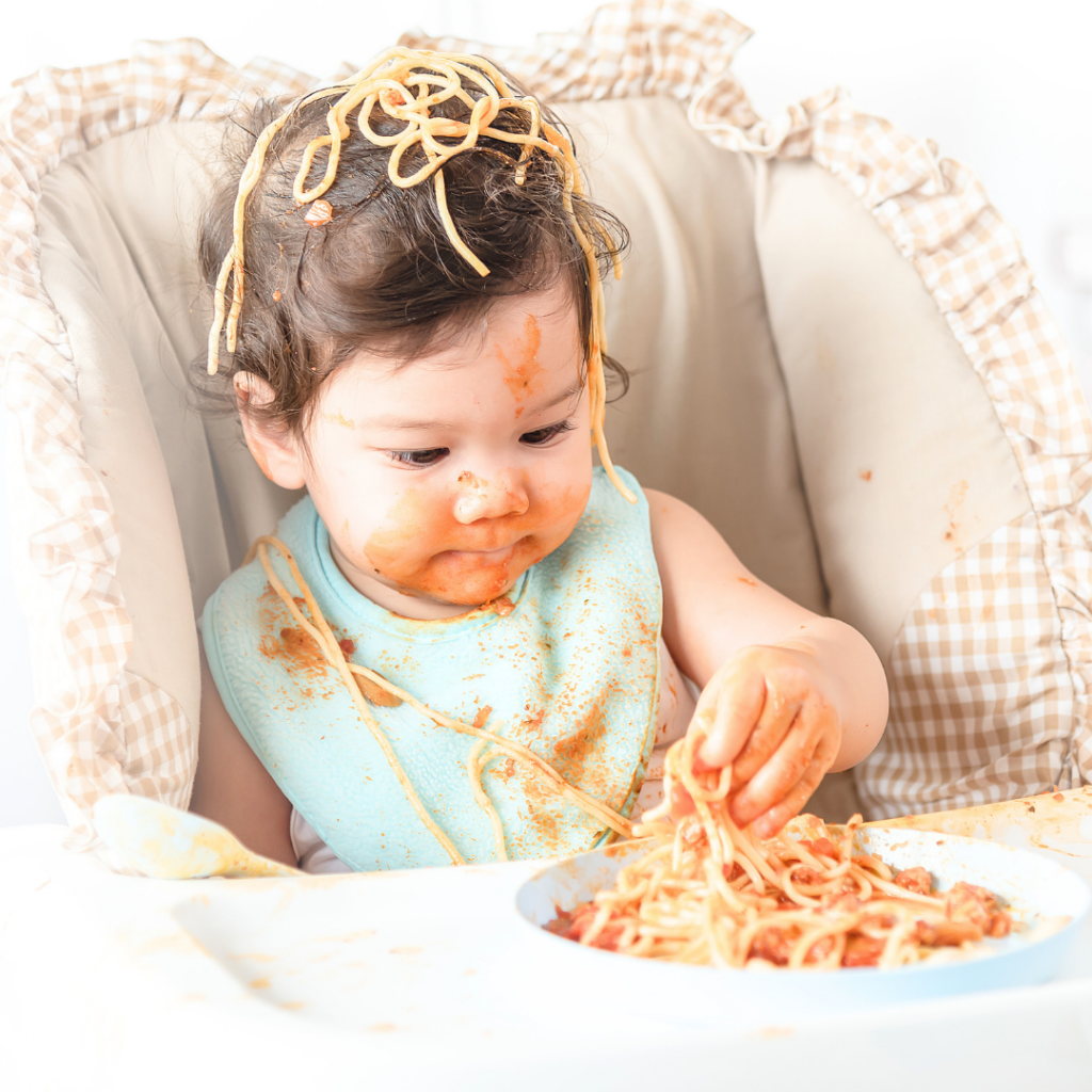 A baby digging their hands into a plate of spaghetti with spaghetti all over their head and face.