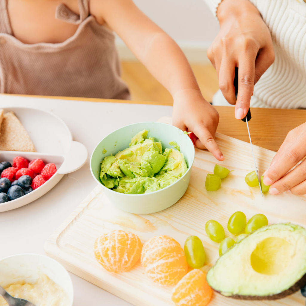 A parent cutting grapes for their baby's breakfast while their baby watches and points towards the grapes, there are also orange slices and mashed avocado in a bowl.