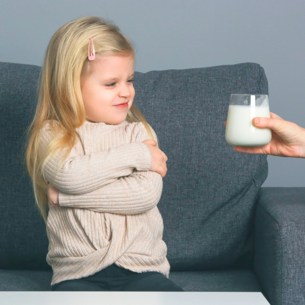 Toddler sitting on the couch with arms crossed, refusing to drink a glass of milk.