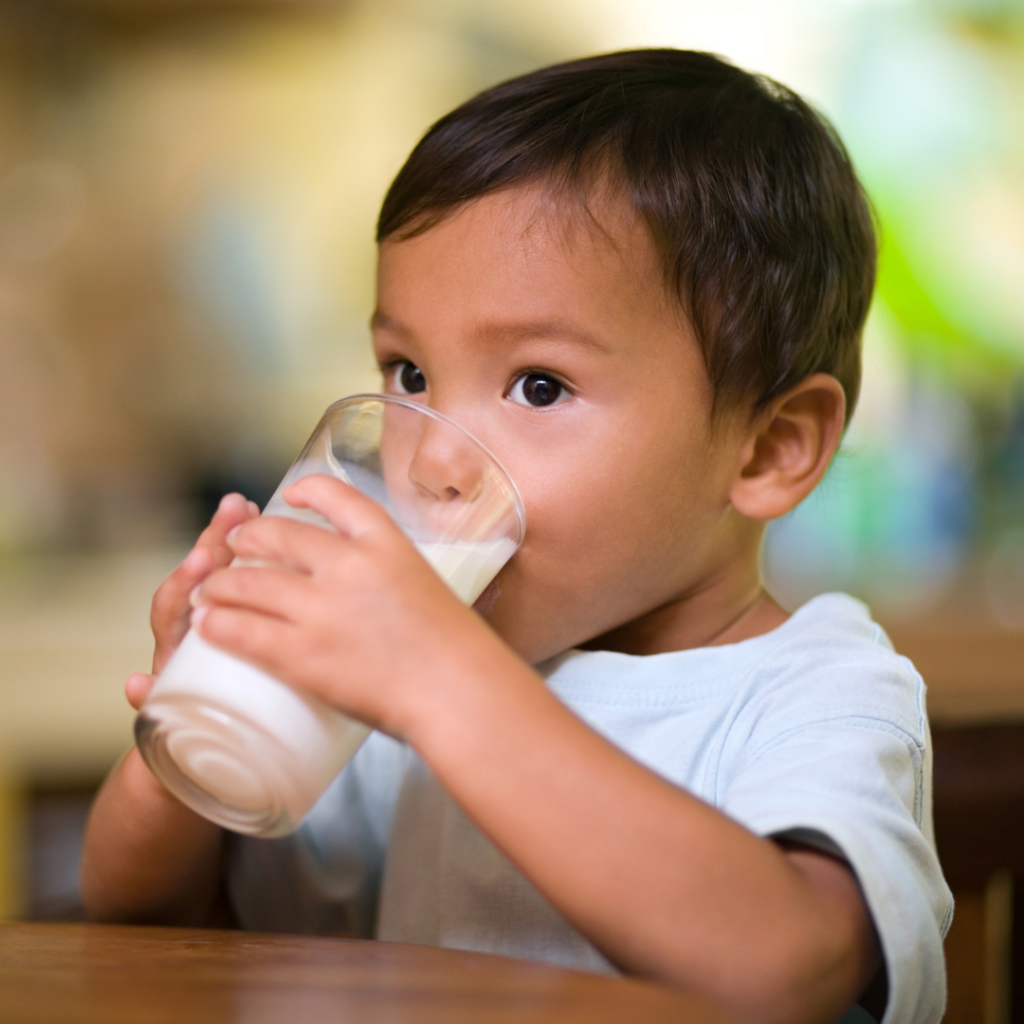 Toddler sitting at the table drinking a tall glass of milk.