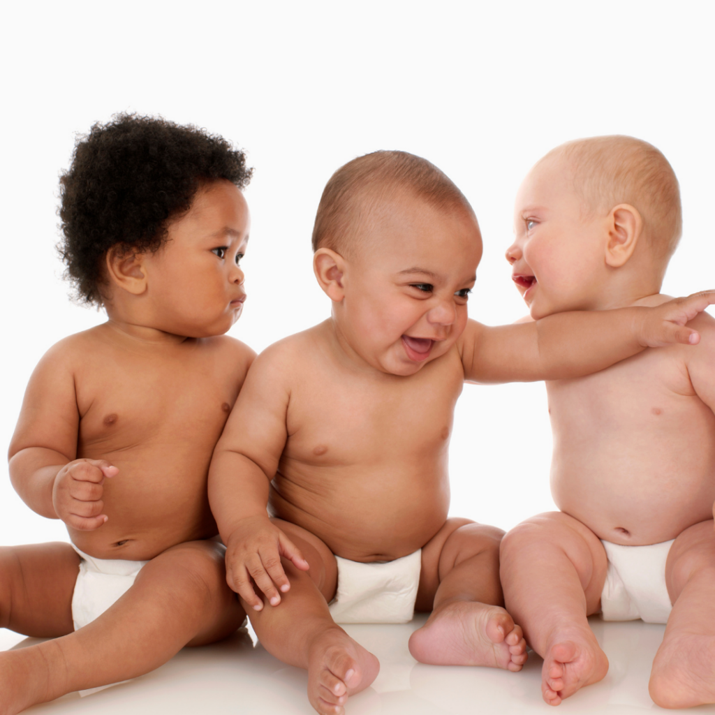 Three babies sitting beside each other, about 6 months old and ready to start weaning, smiling and looking at one another.