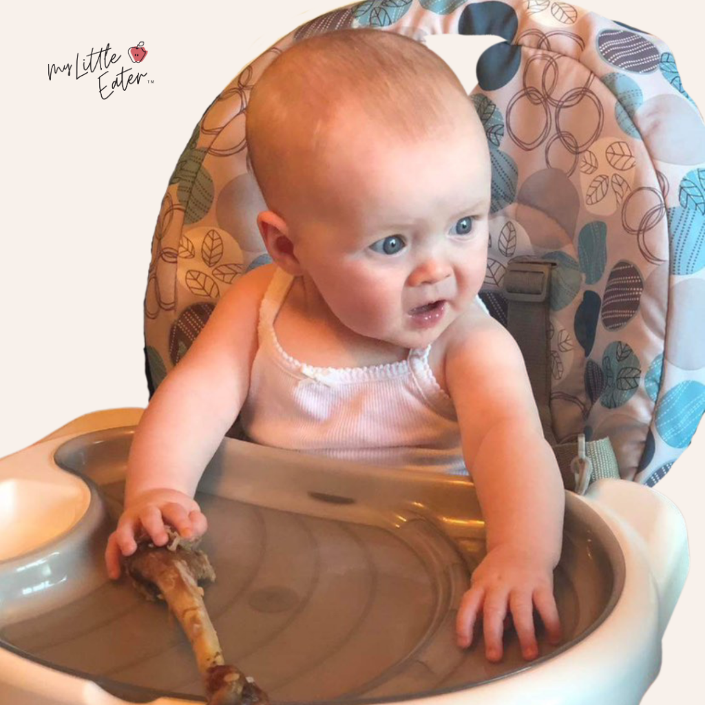 A baby's mouth is open as they sit in their high chair holding a drumstick as a hard munchable for baby led weaning.