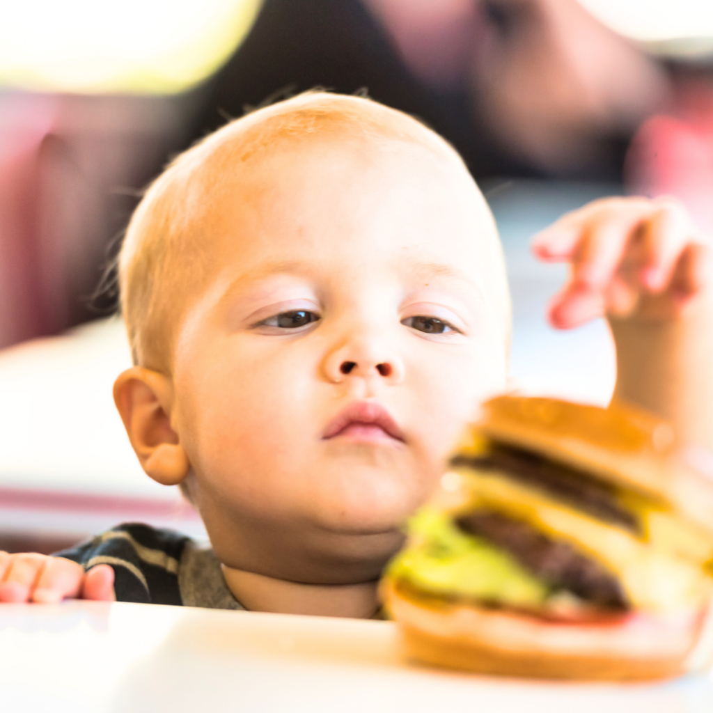 A young toddler reaches for a hamburger, a salty food.