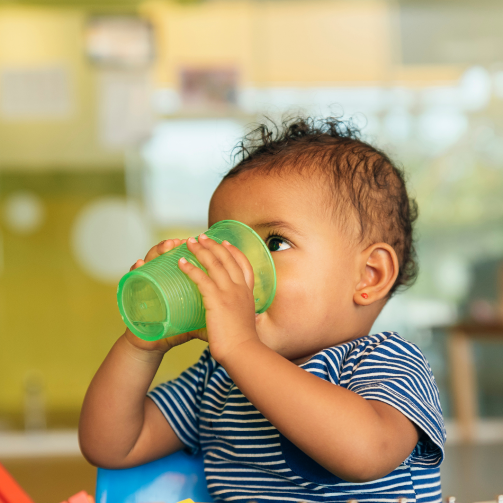 A baby drinks water from an open cup to support baby's digestive system, and keep baby's bowel movements regular by staying hydrated.