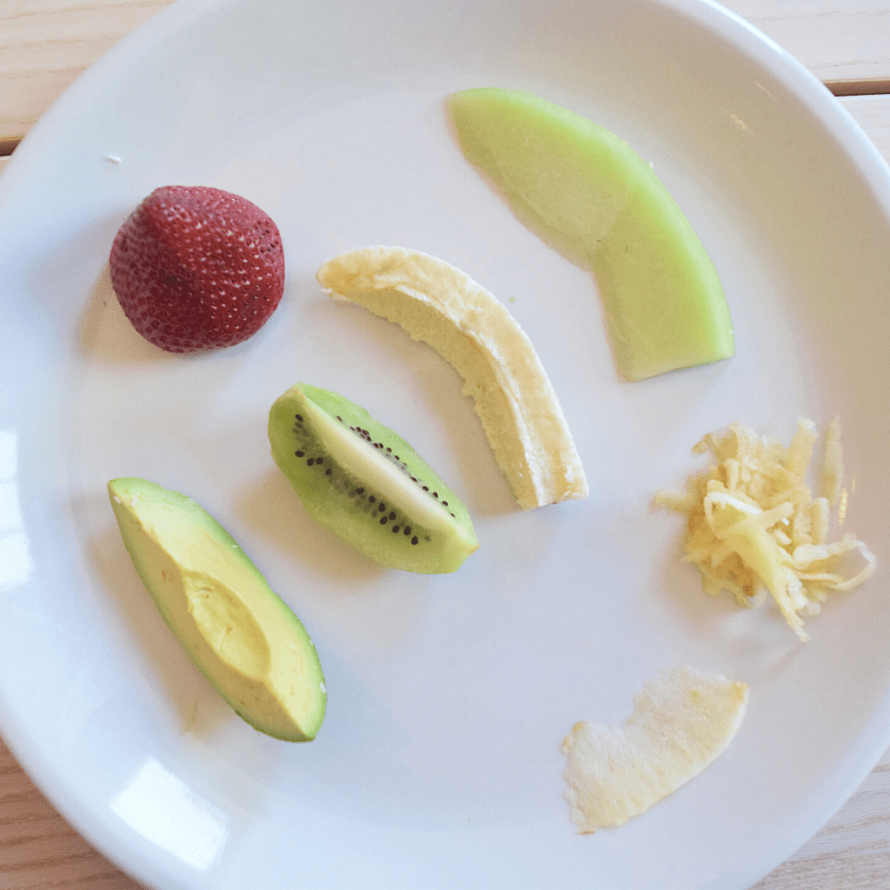 Baby finger foods on a plate ready for a baby starting solid foods, includes a strawberry, avocado wedge, kiwi wedge, banana, shredded apple, and melon.