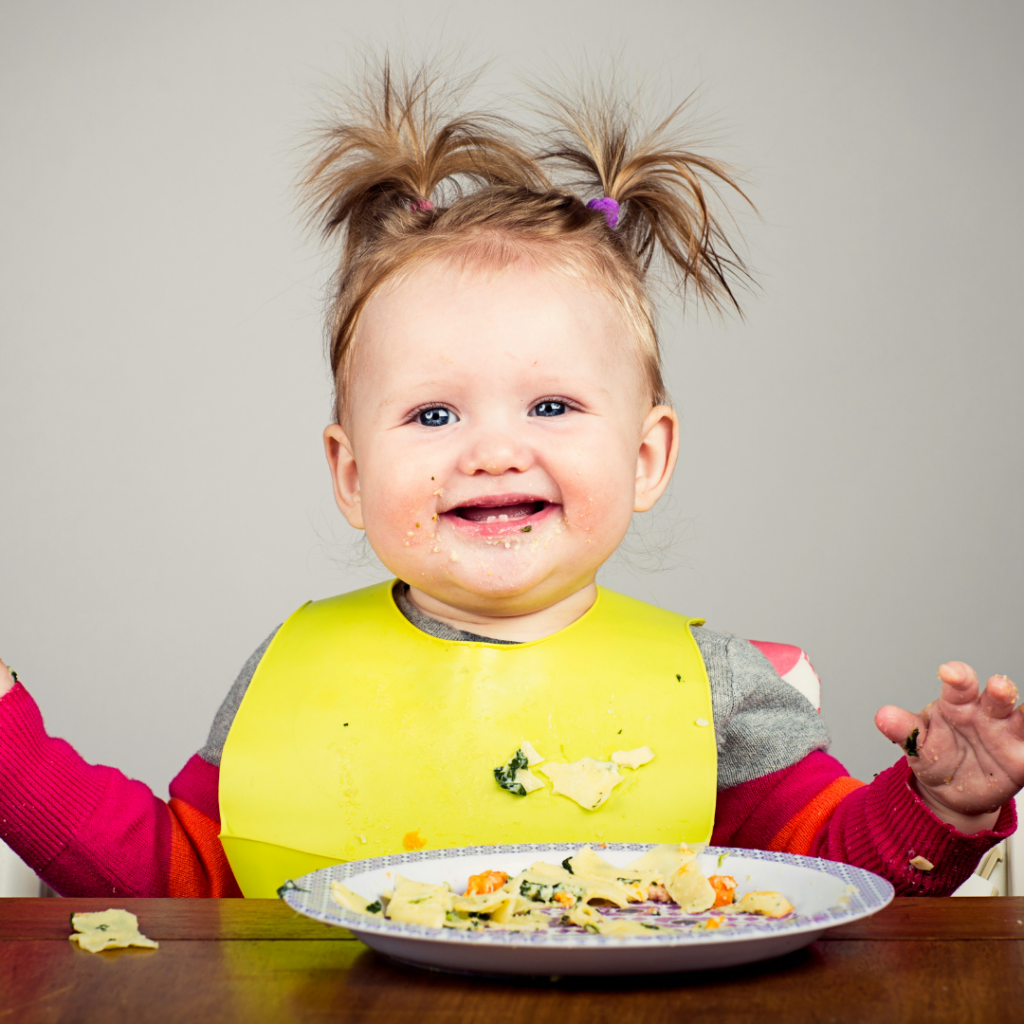 Smiling baby with pigtails sitting at the table eating a meal.