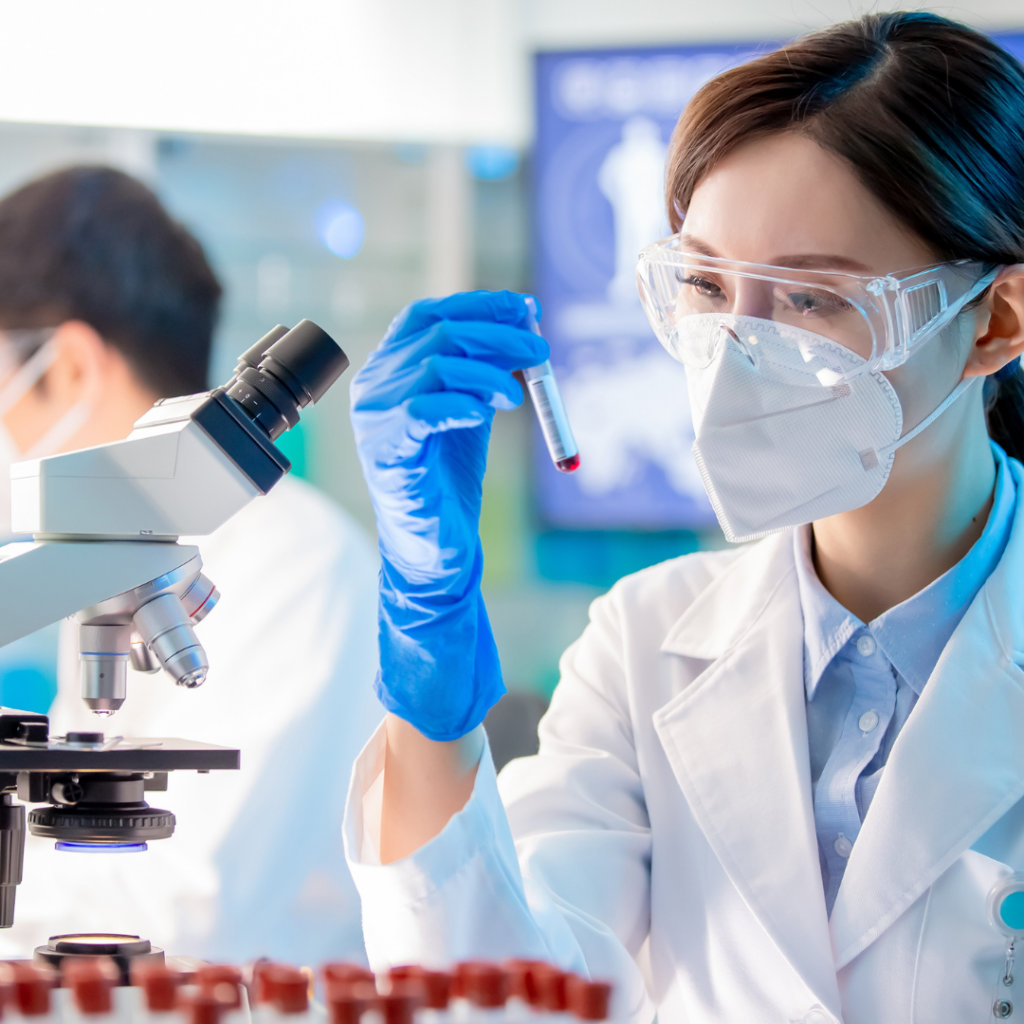 A scientist wearing a mask, eye protection, and rubber gloves examining a test tube.