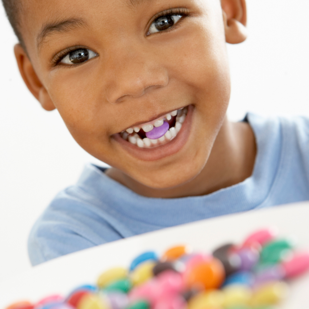 A child smiling with a piece of colored candy in their mouth and a bowl of more candy in front of them.