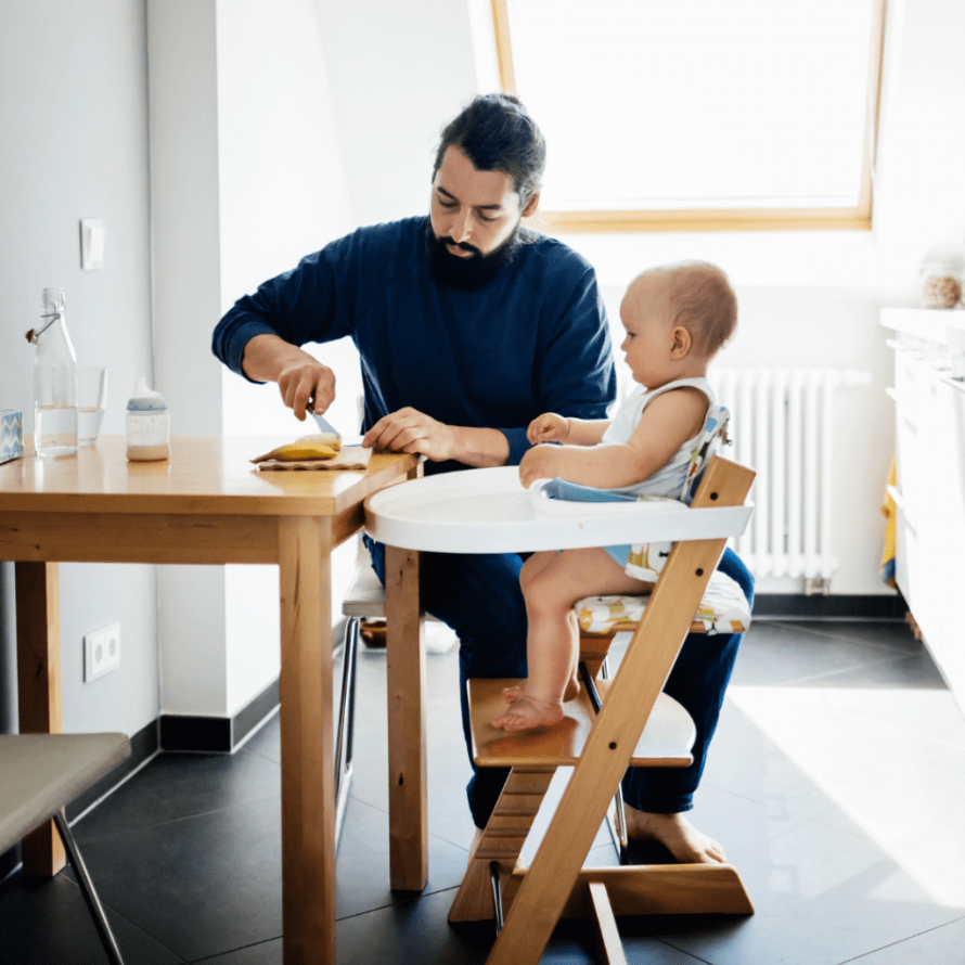 Parent sitting at the table to eat with their baby who is in a high chair.