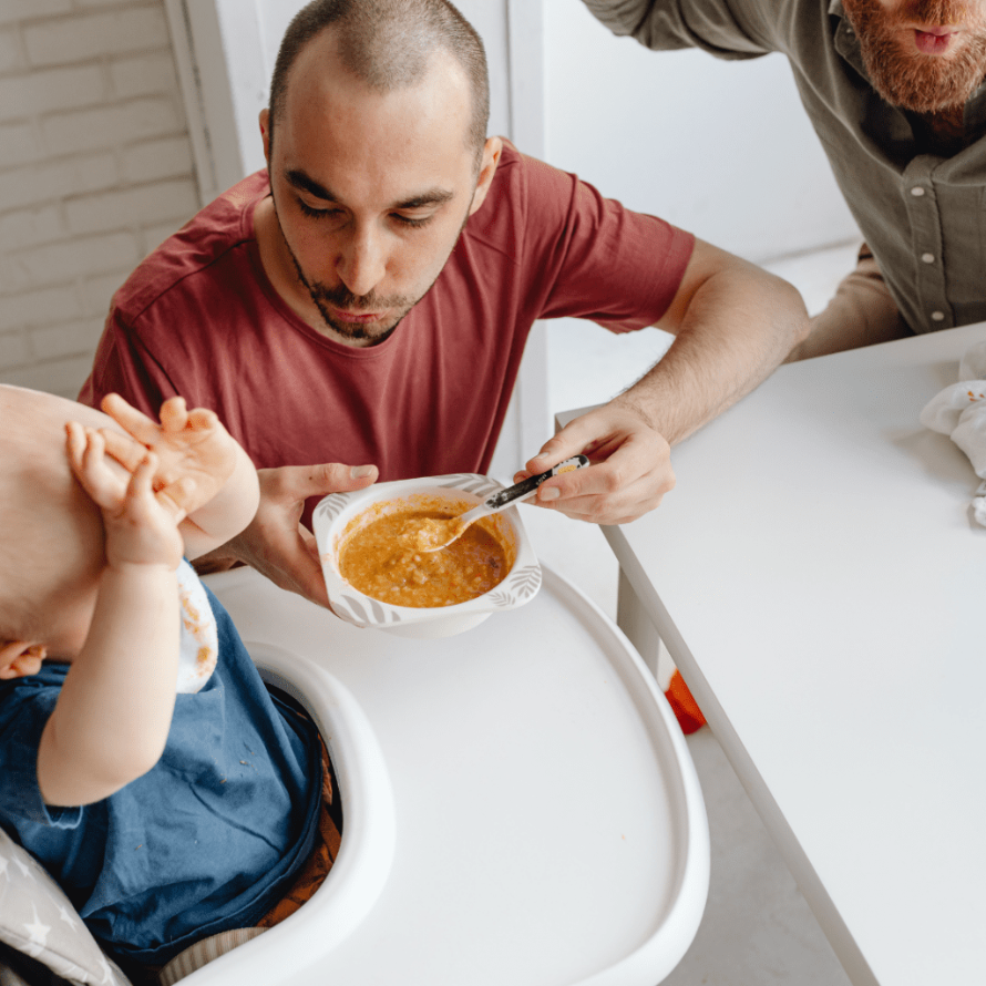 Parents spoon feeding baby while they block their face with their hands.