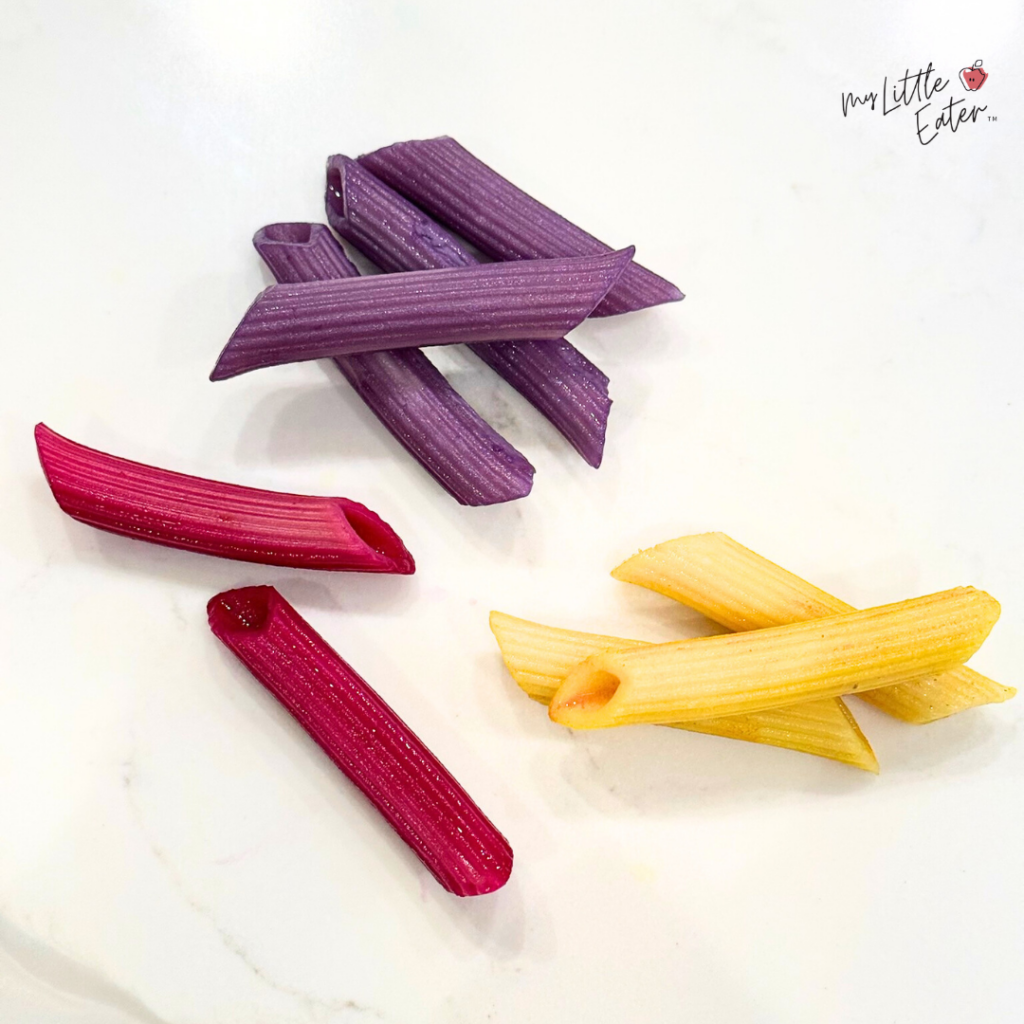 Cooked penne pasta that has been dyed purple, red, and yellow using natural dyes.