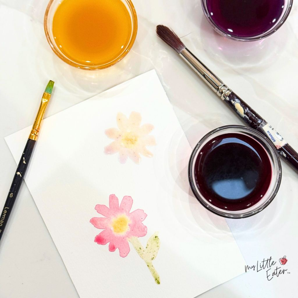 Three natural food colorings in bowls with paintbrushes beside to use the natural food colorings in place of watercolors.