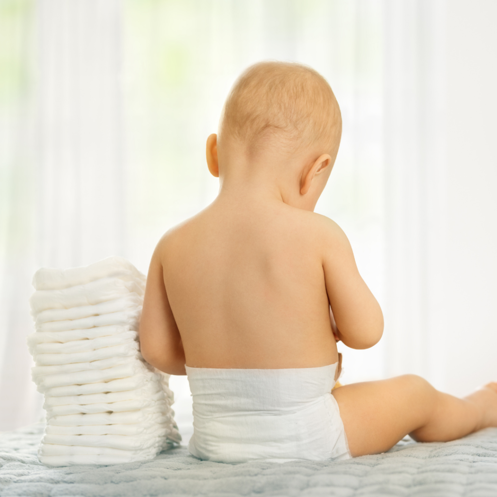 Baby sitting on a bed in just a diaper with a stack of diapers beside them.