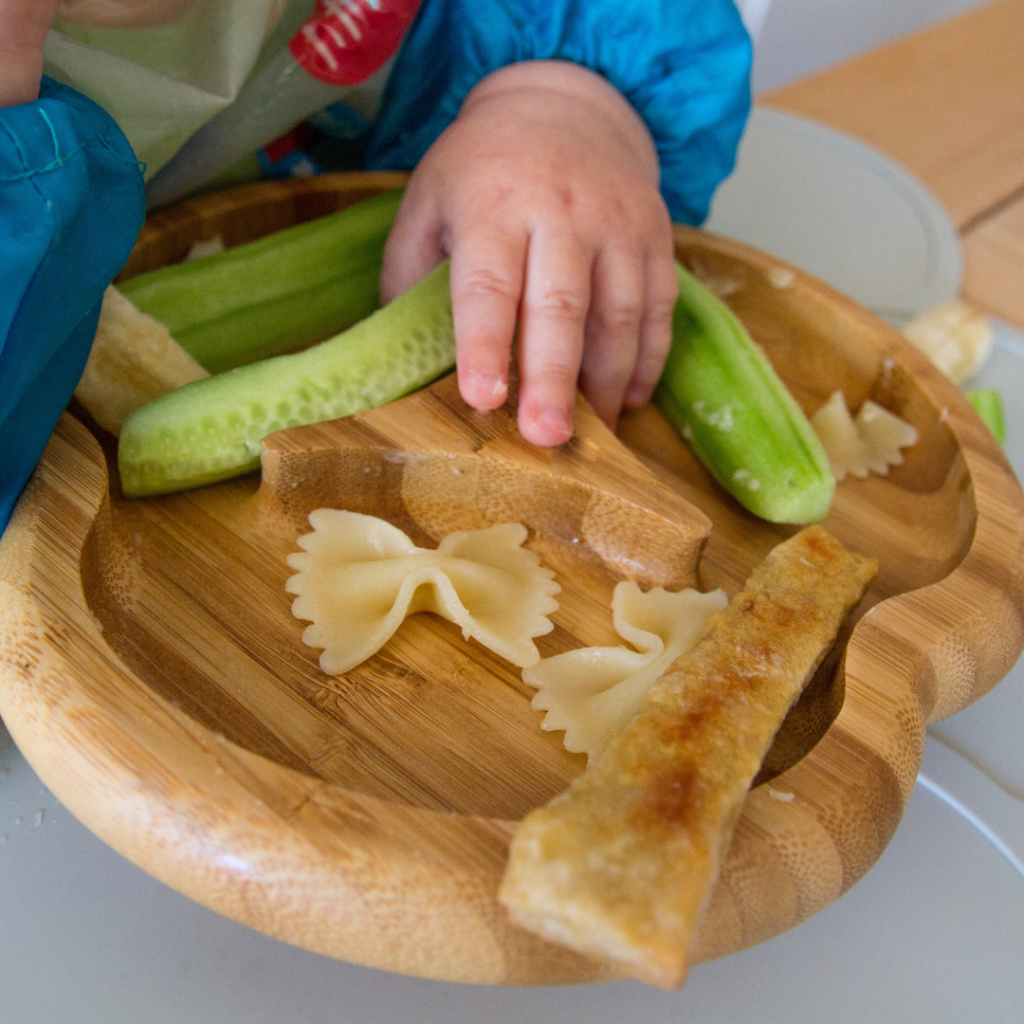 Baby moving their hand across their plate to pick up cooked pasta with their palmar grasp.