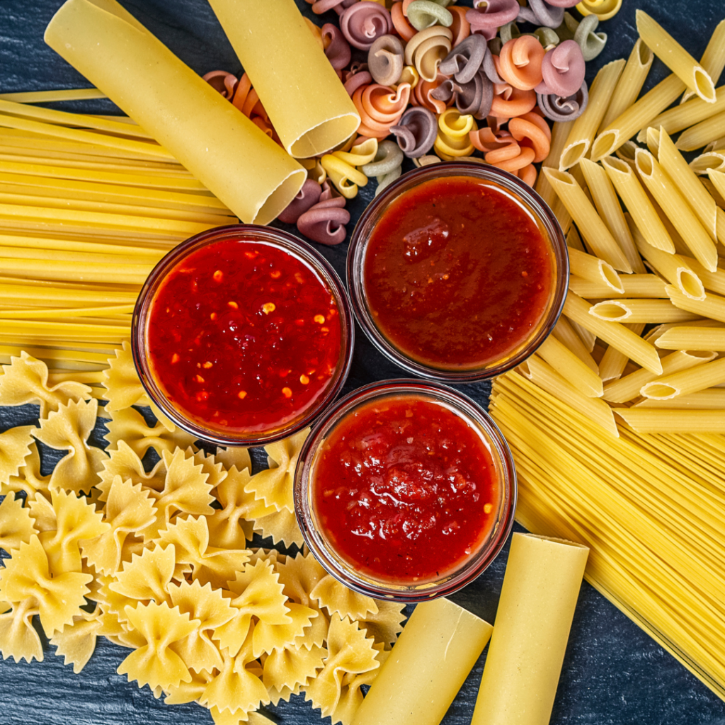 Various shapes of raw pasta spread around 3 small bowls, each containing different sauces.