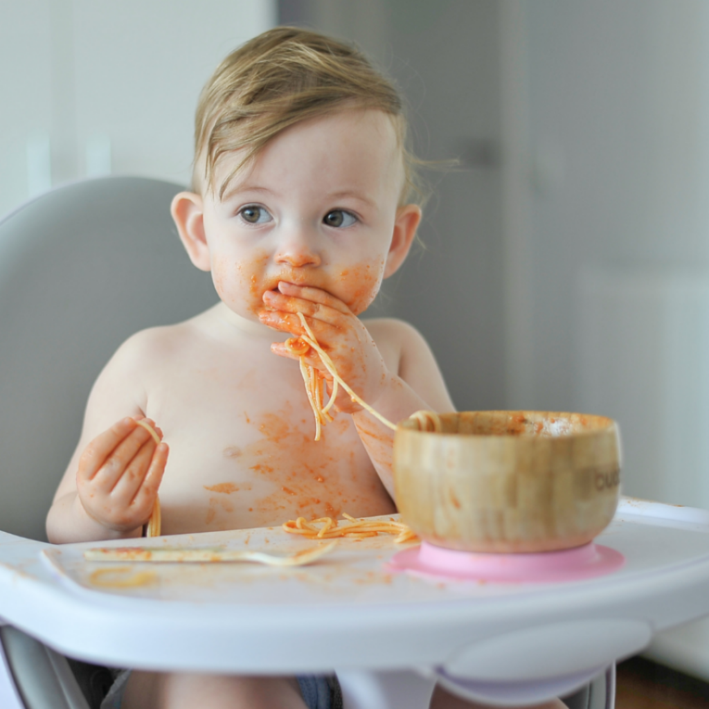 Baby eating spaghetti out of a bowl in their high chair.