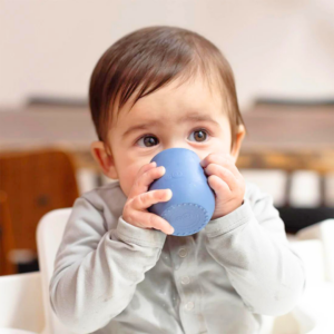 Older baby holds a blue, small open cup for babies, and brings it to baby's lips to drink.