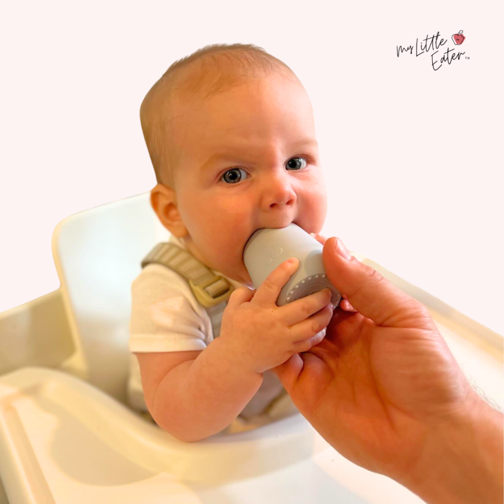 Baby grasping their open cup while the parent still holds on and baby bites the rim of the cup.