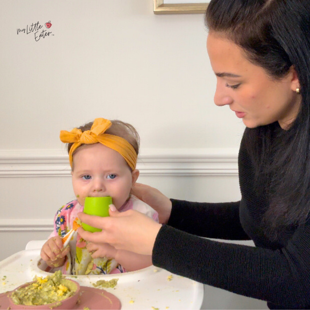 An adult helping a baby drink from a cup by EZPZ during mealtime until she can drink independently.