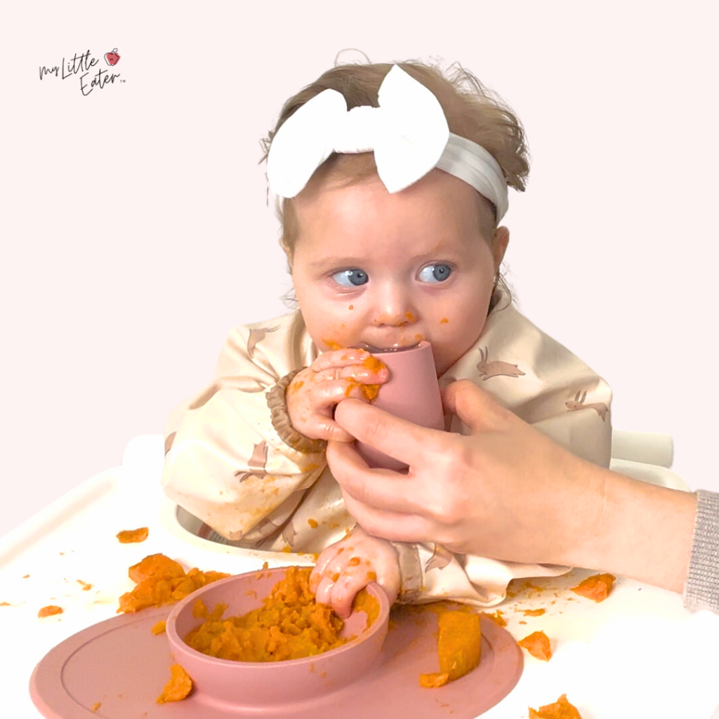 An adult helps their baby drink water from an EZPZ open cup during mealtime.