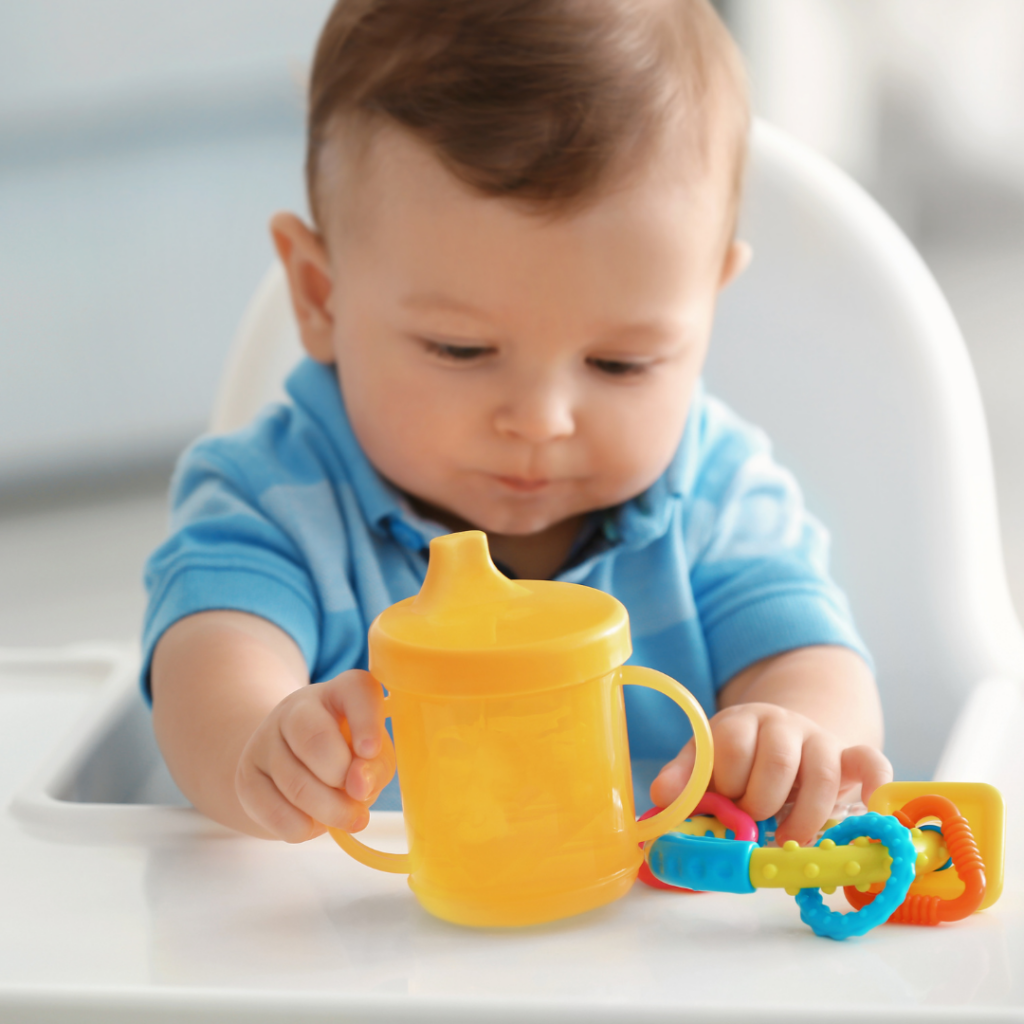 Baby boy sitting in a high chair grabbing the handle of a yellow hard spout sippy cup.