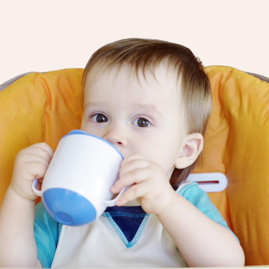 A baby sits in their high chair with an open cup or straw cup to their mouth.