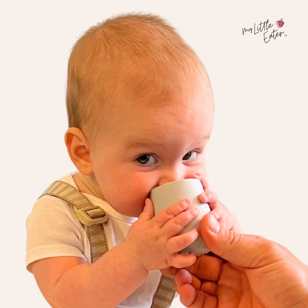 Baby taking a sip of water from an EZPZ open cup with only a small amount of support from Dad’s hand.