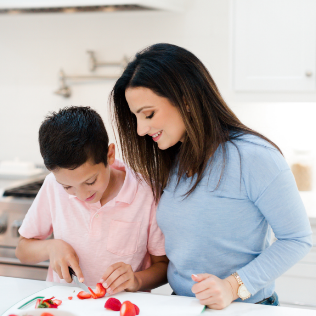 A school-aged child helps their parent with food preparation by slicing strawberries.