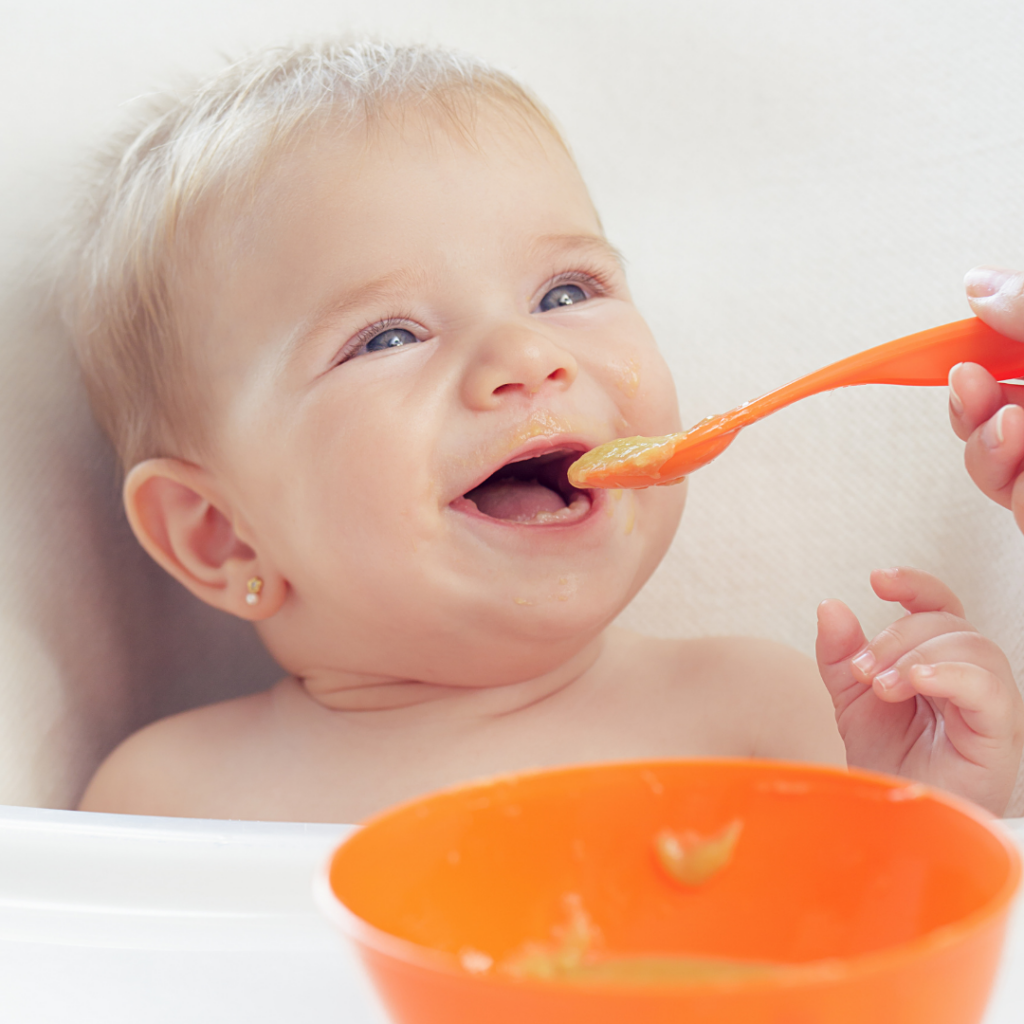 A baby smiling in a high chair, happily being fed a spoon of vegetable puree.