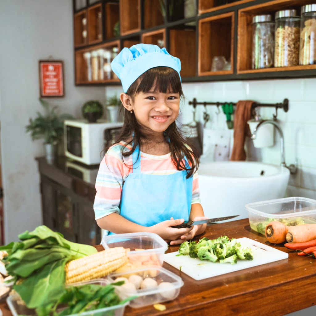 A child, dressed as a chef, chops veggies on their own using sharp knives.