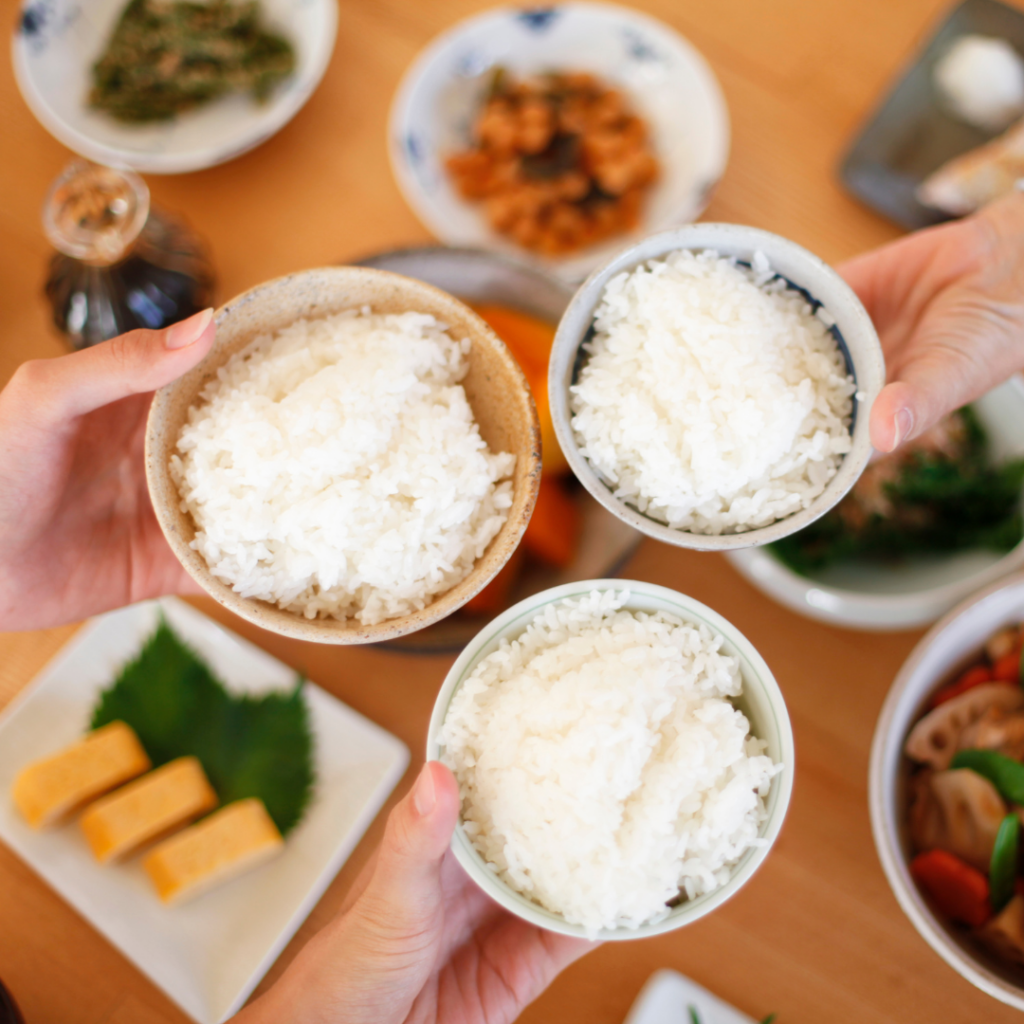 Three people hold a small bowl of rice in their hand with more food in the background.