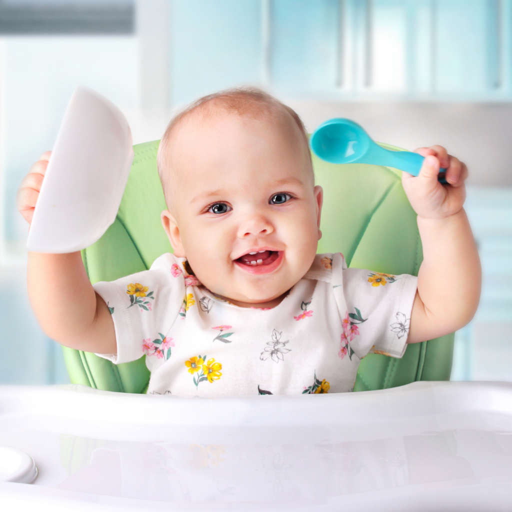 A baby smiled while in their high chair holding a spoon and bowl above their head.