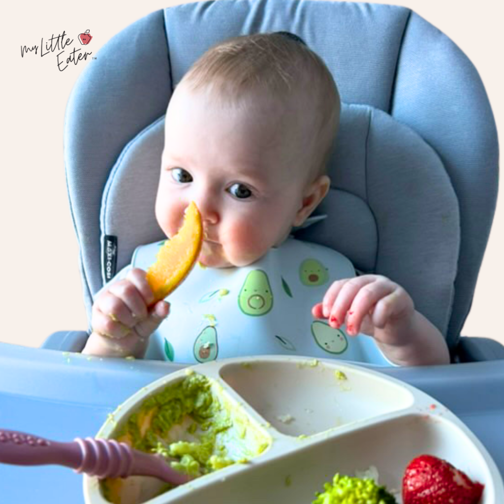 A 6 month old baby in a high chair holding an orange wedge.