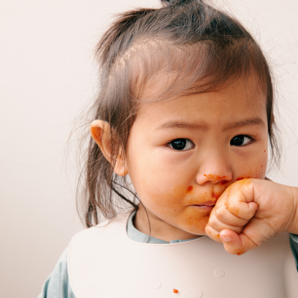 A baby with their hand to their mouth, food on their face, and wearing a bib.