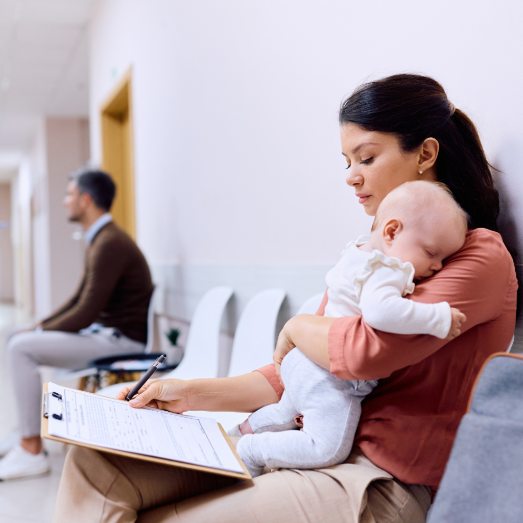 Parent filling out paperwork at a hospital while holding a sleeping baby.