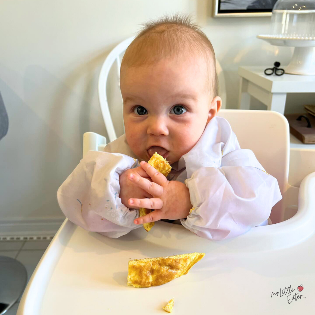 A baby sits in a highchair with a long-sleeved bib on and eats a baby omelet strip.