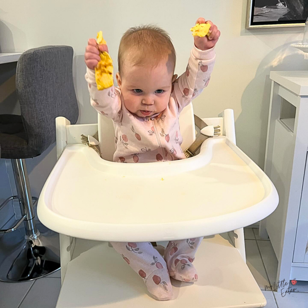 A baby sits in a highchair with a full length pink onesie on and reaches her arms in the air, both hands holding strips of a baby omelet.