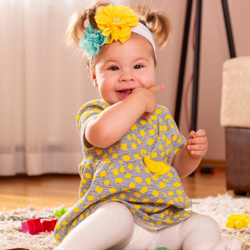 Baby sits on the floor, surrounded by toys, chewing on fingers with the molar area of their mouth.