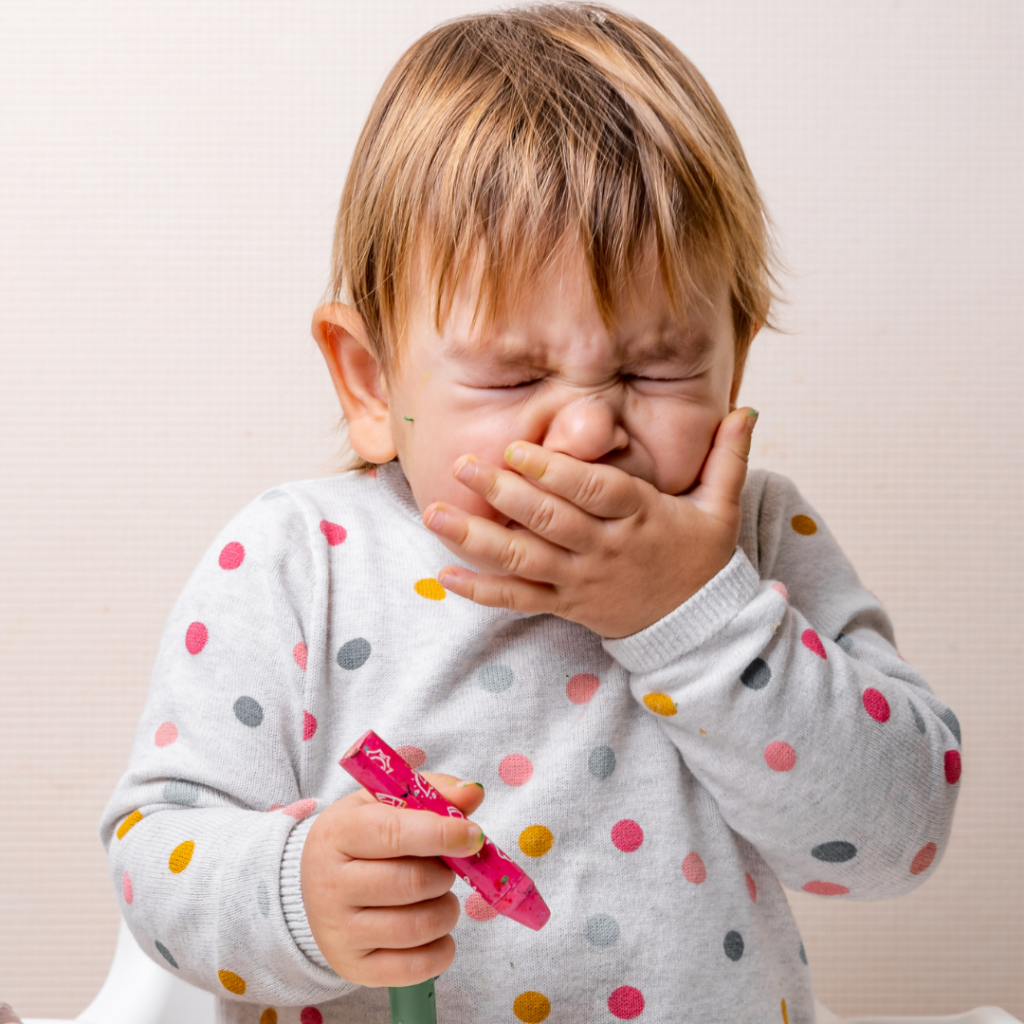 A baby covers their mouth with their hand while they sneeze.