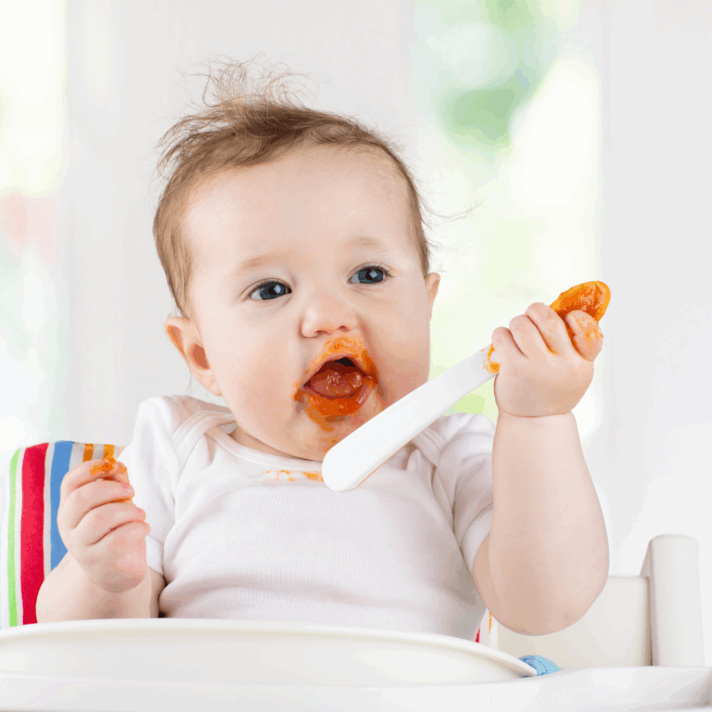 Baby in high chair self-feeding orange puree from a white spoon.