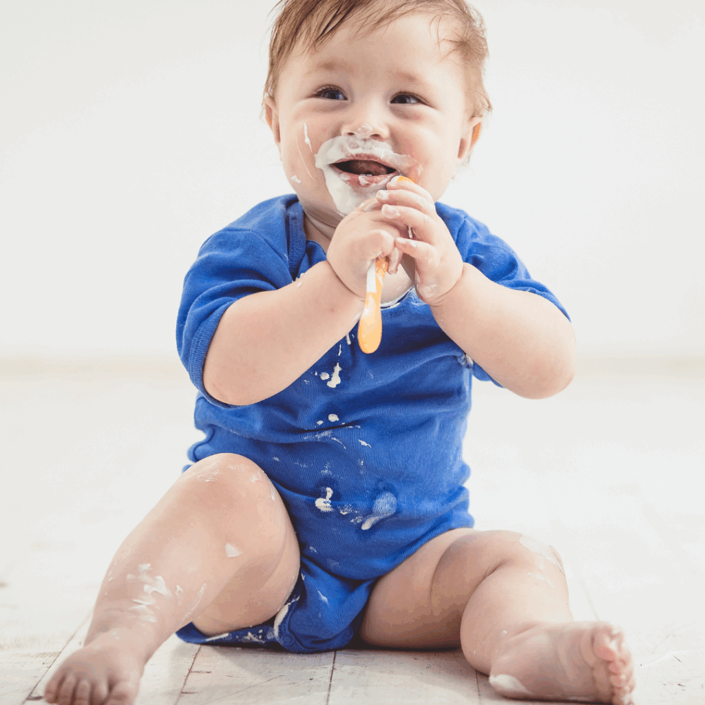 Baby dressed in a blue onesie smiling and looking up, holding a spoon with face, clothes, and legs covered in white yogurt.
