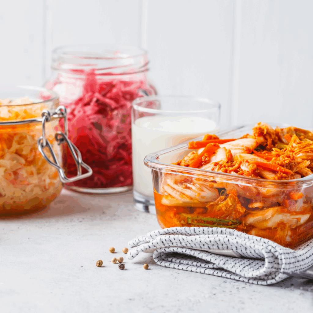 Various fermented foods in glass containers on a light background, including kimchi and a glass of kefir.